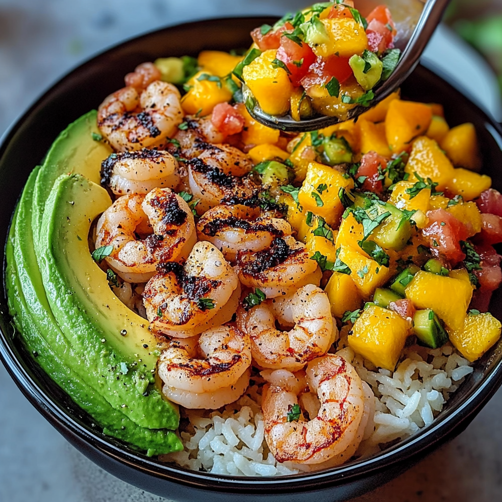 Colorful bowl featuring grilled shrimp, sliced avocado, mango salsa, and lime-chili sauce over rice.
